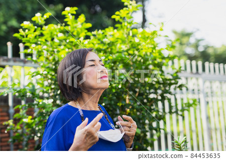 Senior woman holding protective mask in her hand standing in the garden for fresh air. 84453635