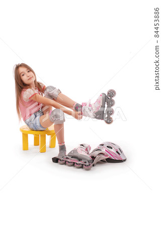 Charming broun-haired girl of school age in short jeans shorts and a pink t-shirt sitting on the chair and tries to foot roller skates. 84453886