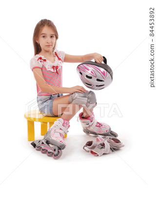 Charming broun-haired girl of school age in short jeans shorts and a pink t-shirt sitting on the chair and tries to foot roller skates. 84453892