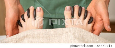 Feet massage with black pebbles between the female toes in hands of the masseur in the spa. Feet massage with black pebbles between the female toes in hands of the masseur in the spa. 84460713