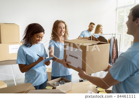 Millennial volunteer giving carton box with donations to woman, female social worker making notes in clipboard Millennial volunteer giving carton box with donations to woman, female social worker making notes in clipboard 84461111