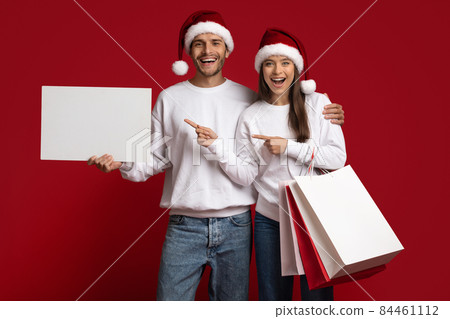 Couple Wearing Santa Hats Holding Shopping Bags And Pointing At Blank Placard Couple Wearing Santa Hats Holding Shopping Bags And Pointing At Blank Placard 84461112