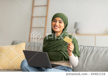 Beautiful young Arab woman in hijab using credit card and laptop, sitting on couch, buying goods online from home Beautiful young Arab woman in hijab using credit card and laptop, sitting on couch, buying goods online from home 84461316