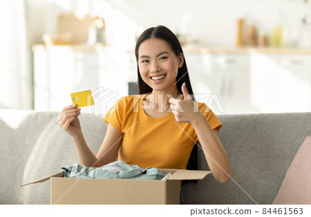 Home delivery. Young asian woman sitting on sofa with parcel, showing thumb up and credit card, smiling at camera 84461563