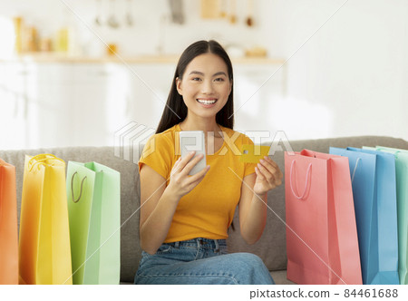 Positive asian lady with credit card and smartphone sitting on sofa surrounded by colorful shopping bags Positive asian lady with credit card and smartphone sitting on sofa surrounded by colorful shopping bags 84461688