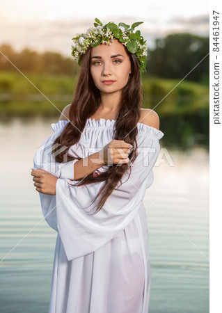 Beautiful black haired girl in white vintage dress and wreath of flowers standing in water of lake. Sun flare. Beautiful black haired girl in white vintage dress and wreath of flowers standing in water of lake. Sun flare. 84461947