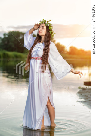 Beautiful black haired girl in white vintage dress and wreath of flowers standing in water of lake. Sun flare. Beautiful black haired girl in white vintage dress and wreath of flowers standing in water of lake. Sun flare. 84461953