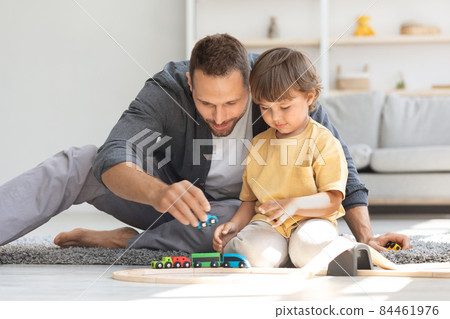 Family fun and leisure. Cute little boy playing wooden railway and trains with his daddy, sitting on floor at home Family fun and leisure. Cute little boy playing wooden railway and trains with his daddy, sitting on floor at home 84461976