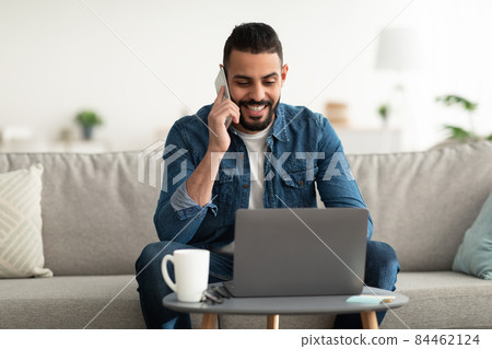 Work from home. Positive young Arab guy talking on mobile phone while using laptop computer indoors 84462124