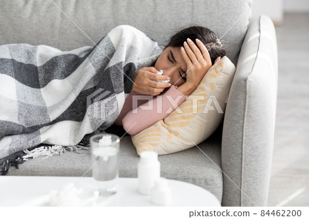Weak sad young hindu lady with plaid, suffers from flu, blows nose in napkin, lies on sofa in living room interior Weak sad young hindu lady with plaid, suffers from flu, blows nose in napkin, lies on sofa in living room interior 84462200