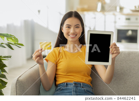Positive asian woman showing blank tablet screen and credit card, sitting on sofa and smiling, mockup 84462241