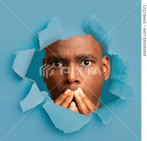 Surprised young african american guy with wide open eyes closed his mouth with hands, looks through hole in blue paper Surprised young african american guy with wide open eyes closed his mouth with hands, looks through hole in blue paper 84462321