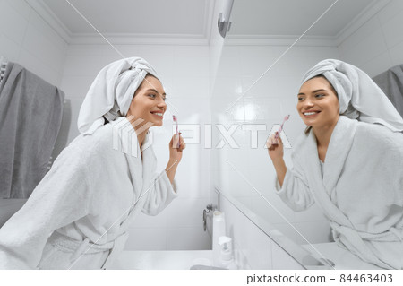 Pretty woman in white robe with towel on head brush teeth in bathroom. 84463403