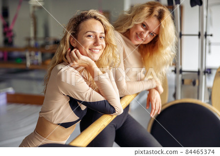 Portrait of smiling young woman looking at camera while standing near ballet barre after pilates workout together with her instructor in stretching studio 84465374