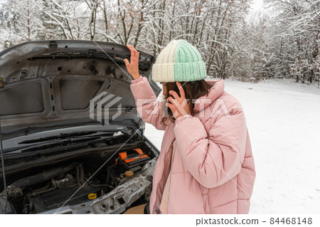 Young brunette woman in a pink winter down jacket and warm hat is standing near a car with an open hood and talking on the phone Young brunette woman in a pink winter down jacket and warm hat is standing near a car with an open hood and talking on the phone 84468148