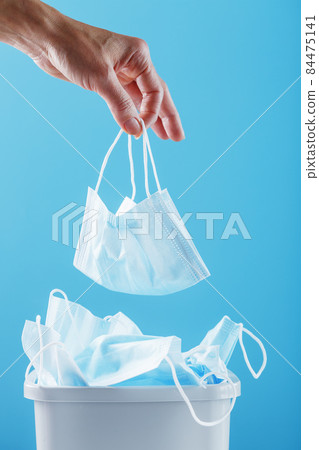 A hand throws a used protective mask into a trash can filled with masks from a virus on a blue background. A hand throws a used protective mask into a trash can filled with masks from a virus on a blue background. 84475141