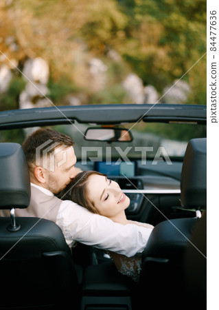 Groom hugs bride who has laid her head on his shoulder in a convertible Groom hugs bride who has laid her head on his shoulder in a convertible 84477636