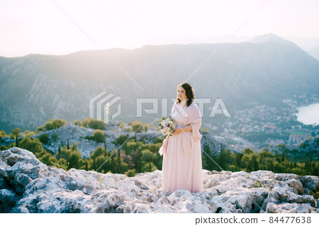 Bride with a bouquet stands on the stones against the background of the mountains Bride with a bouquet stands on the stones against the background of the mountains 84477638