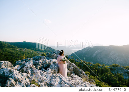 Bride with a bouquet stands on the stones and looks at the mountains Bride with a bouquet stands on the stones and looks at the mountains 84477640