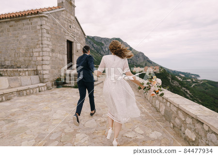 Groom and bride with a bouquet of flowers run to the church on the mountain. Back view Groom and bride with a bouquet of flowers run to the church on the mountain. Back view 84477934