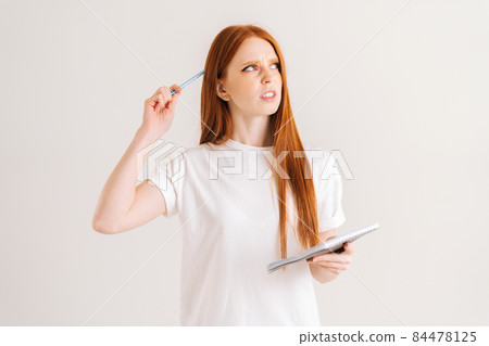 Studio portrait of confused redhead young woman scratches head with pencil, holding diary looking down, standing on white isolated background. Studio portrait of confused redhead young woman scratches head with pencil, holding diary looking down, standing on white isolated background. 84478125