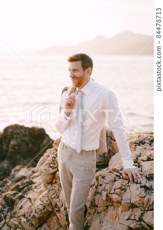 Groom with a jacket on his shoulder stands on a rock near the sea Groom with a jacket on his shoulder stands on a rock near the sea 84478713