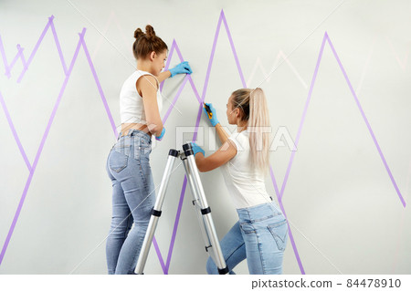two Young women preparing wall for painting standing on ladder applying masking tape on wall. Diy project two Young women preparing wall for painting standing on ladder applying masking tape on wall. Diy project 84478910