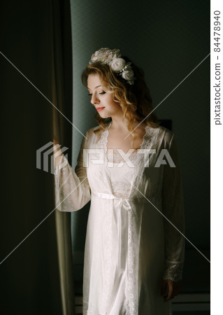 A bride in a white wreath and lace peignoir stands at the window and peeps out from behind the curtain in a hotel room A bride in a white wreath and lace peignoir stands at the window and peeps out from behind the curtain in a hotel room 84478940