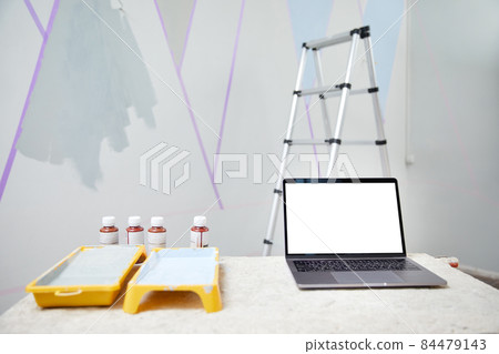 Laptop standing on table with white copy space mockup with wall being painted on background. Ladder and paint. Renovation and home decor concept Laptop standing on table with white copy space mockup with wall being painted on background. Ladder and paint. Renovation and home decor concept 84479143