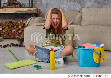 Frustrated young woman sitting on living room floor with cleaning supplies and equipment and screaming 84484116