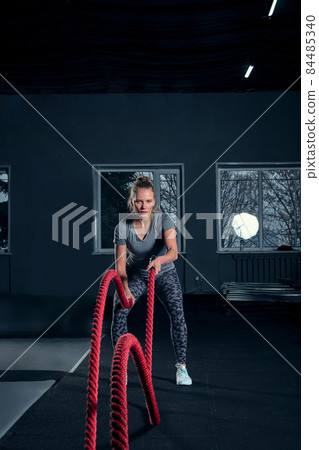 Vertical full length shot of an athletic fitness woman doing functional training exercise with battle ropes at the gym Cross Fit cross training strength endurance energy power concept Vertical full length shot of an athletic fitness woman doing functional training exercise with battle ropes at the gym Cross Fit cross training strength endurance energy power concept 84485340