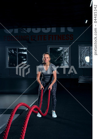 Vertical full length shot of an athletic fitness woman doing functional training exercise with battle ropes at the gym CrossFit cross training strength endurance energy power concept 84485342