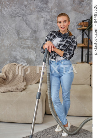 Young smiling woman vacuum cleaning the carpet in the living room, modern scandinavian interior. Home, housekeeping concept Young smiling woman vacuum cleaning the carpet in the living room, modern scandinavian interior. Home, housekeeping concept 84485526