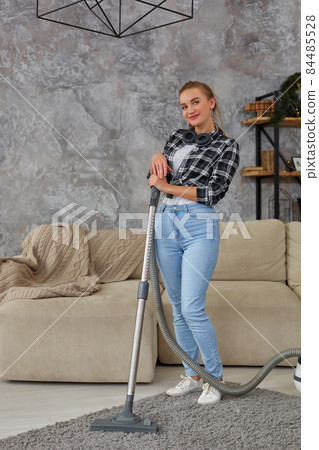 Young smiling woman vacuum cleaning the carpet in the living room, modern scandinavian interior. Home, housekeeping concept Young smiling woman vacuum cleaning the carpet in the living room, modern scandinavian interior. Home, housekeeping concept 84485528