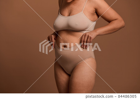 Cropped view of a woman in beige underwear laying her hands on her stomach with relaxed muscles that have lost their tone and stretch marks after pregnancy and childbirth. Body positive concept 84486305