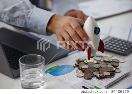 Person hold rocket model over bunch of coins and business papers on table 84486379