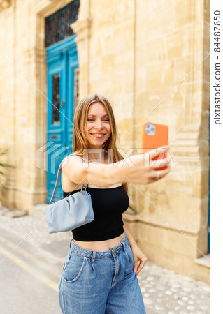 Young beautiful girl in denim jeans and purse handbag makes selfie against the background of the Cyprus old town. Young beautiful girl in denim jeans and purse handbag makes selfie against the background of the Cyprus old town. 84487850