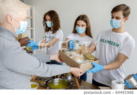 Group of young volunteers in masks giving food in carton boxes as donation for poor and elderly people Group of young volunteers in masks giving food in carton boxes as donation for poor and elderly people 84491986