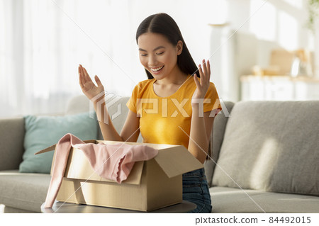 Unpacking clothes. Portrait of happy asian lady opening cardboard box, taking out new shirt, sitting on sofa at home Unpacking clothes. Portrait of happy asian lady opening cardboard box, taking out new shirt, sitting on sofa at home 84492015