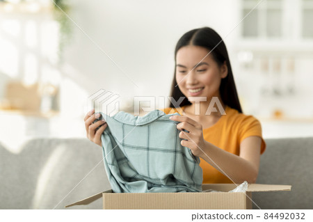 Buying clothes via internet. Happy young asian lady unboxing cardboard delivery package after online shopping Buying clothes via internet. Happy young asian lady unboxing cardboard delivery package after online shopping 84492032