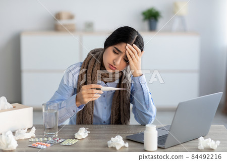 Sad young hindu woman manager suffering from headache, colds and flu looks at thermometer Sad young hindu woman manager suffering from headache, colds and flu looks at thermometer 84492216