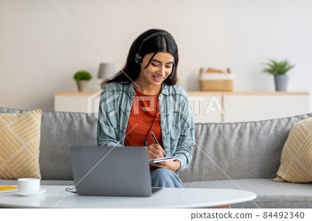 Young Arab woman in headphones using laptop, working or studying online from home, taking notes during business meeting Young Arab woman in headphones using laptop, working or studying online from home, taking notes during business meeting 84492340