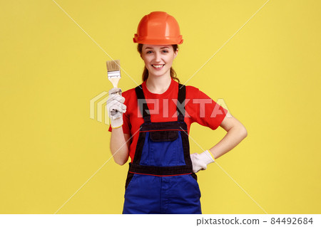 Positive young adult builder woman standing with paintbrush in hands, wearing work uniform and protective helmet, keeps hand on waist. Indoor studio shot isolated on yellow background. Positive young adult builder woman standing with paintbrush in hands, wearing work uniform and protective helmet, keeps hand on waist. Indoor studio shot isolated on yellow background. 84492684