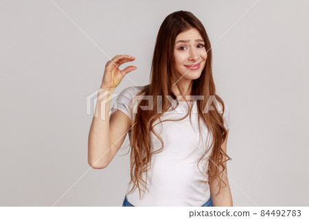 Portrait of unhappy woman with dark hair showing little size gesture with disappointed frustrated face expression, wearing white T-shirt. Indoor studio shot isolated on gray background. Portrait of unhappy woman with dark hair showing little size gesture with disappointed frustrated face expression, wearing white T-shirt. Indoor studio shot isolated on gray background. 84492783