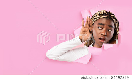Surprised interested millennial black female with open mouth puts her hand to ear and listens through hole in pink paper 84492819