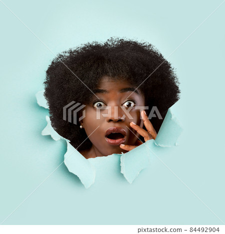 Amazed surprised millennial african american woman with open mouth looking through hole in blue paper Amazed surprised millennial african american woman with open mouth looking through hole in blue paper 84492904