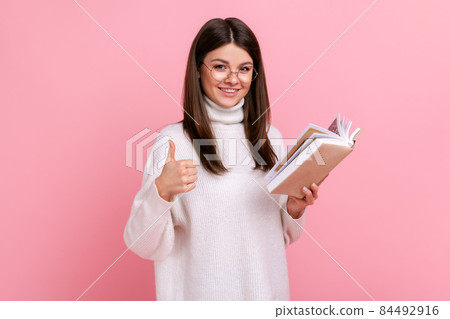 Beautiful satisfied woman showing thumbs up gesture holding and reading book, likes genre and plot, wearing white casual style sweater. Indoor studio shot isolated on pink background. Beautiful satisfied woman showing thumbs up gesture holding and reading book, likes genre and plot, wearing white casual style sweater. Indoor studio shot isolated on pink background. 84492916