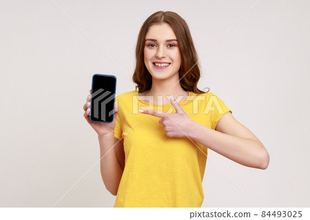 Look at my cellphone. Adorable glad teenager girl pointing smartphone with finger and smiling at camera, advertising of mobile device. Indoor studio shot isolated on gray background. Look at my cellphone. Adorable glad teenager girl pointing smartphone with finger and smiling at camera, advertising of mobile device. Indoor studio shot isolated on gray background. 84493025