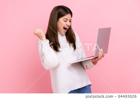 Satisfied female standing with laptop and clenching her fist, celebrating success, yelling happily, wearing white casual style sweater. Indoor studio shot isolated on pink background. Satisfied female standing with laptop and clenching her fist, celebrating success, yelling happily, wearing white casual style sweater. Indoor studio shot isolated on pink background. 84493026