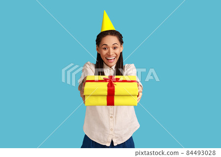 Happy excited woman with black dreadlocks standing in part cone and giving present box, celebrating friend's birthday, wearing white shirt. Indoor studio shot isolated on blue background. Happy excited woman with black dreadlocks standing in part cone and giving present box, celebrating friend's birthday, wearing white shirt. Indoor studio shot isolated on blue background. 84493028
